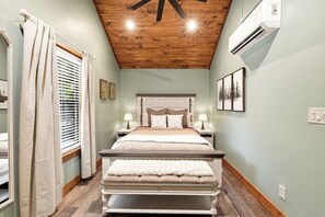 Un dormitorio con techo de madera, una cama con cabecera, mesitas de noche con lámparas y una ventana con cortinas.