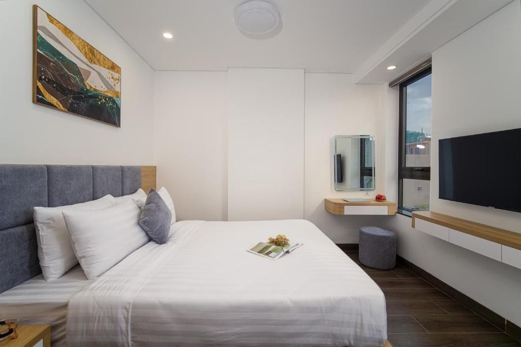 A modern hotel room with a large bed, a bedside table, a small stool, a TV, and a window.
