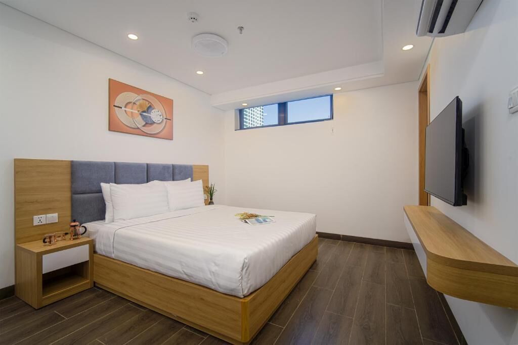 A modern bedroom with a wooden bed frame, a nightstand, a flat-screen TV mounted on the wall, and a window providing natural light.