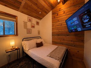 Ein Schlafzimmer mit einem Bett, einer Nachttischlampe, einem an der Wand montierten Fernseher und einem Fenster mit Blick auf Bäume.