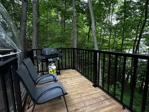 Eine Holzterrasse mit Grill und St ühlen, umgeben von einem schwarzen Geländer und dichtem Wald.