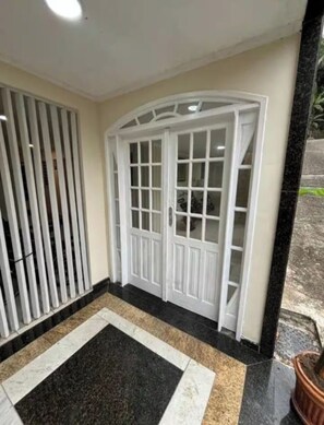 Une porte blanche avec des panneaux de verre, un hall d’entrée carrelé en noir et beige, et un portail métallique sur la gauche.