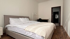 Cama bem arrumada com cabeceira, mesa de cabeceira, luminária e porta que dá para outro quarto.