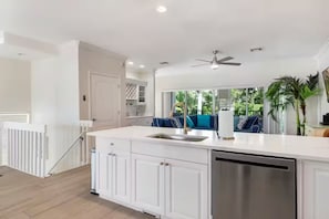 A modern kitchen with white cabinetry, a stainless steel dishwasher, and a large window overlooking a balcony with blue outdoor seating. - NEW! Steps to Beach, Hot Tub, Pool, Kid Friendly (Holmes Beach)
