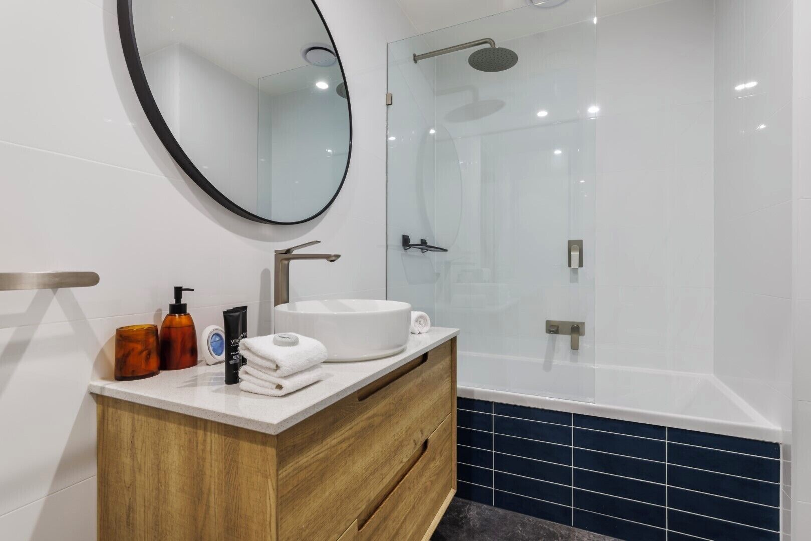 A modern bathroom with a round mirror, a wooden vanity, a white sink, and a glass-enclosed shower.