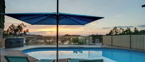 A swimming pool with lounge chairs and an umbrella, surrounded by a fence and a view of the sunset.