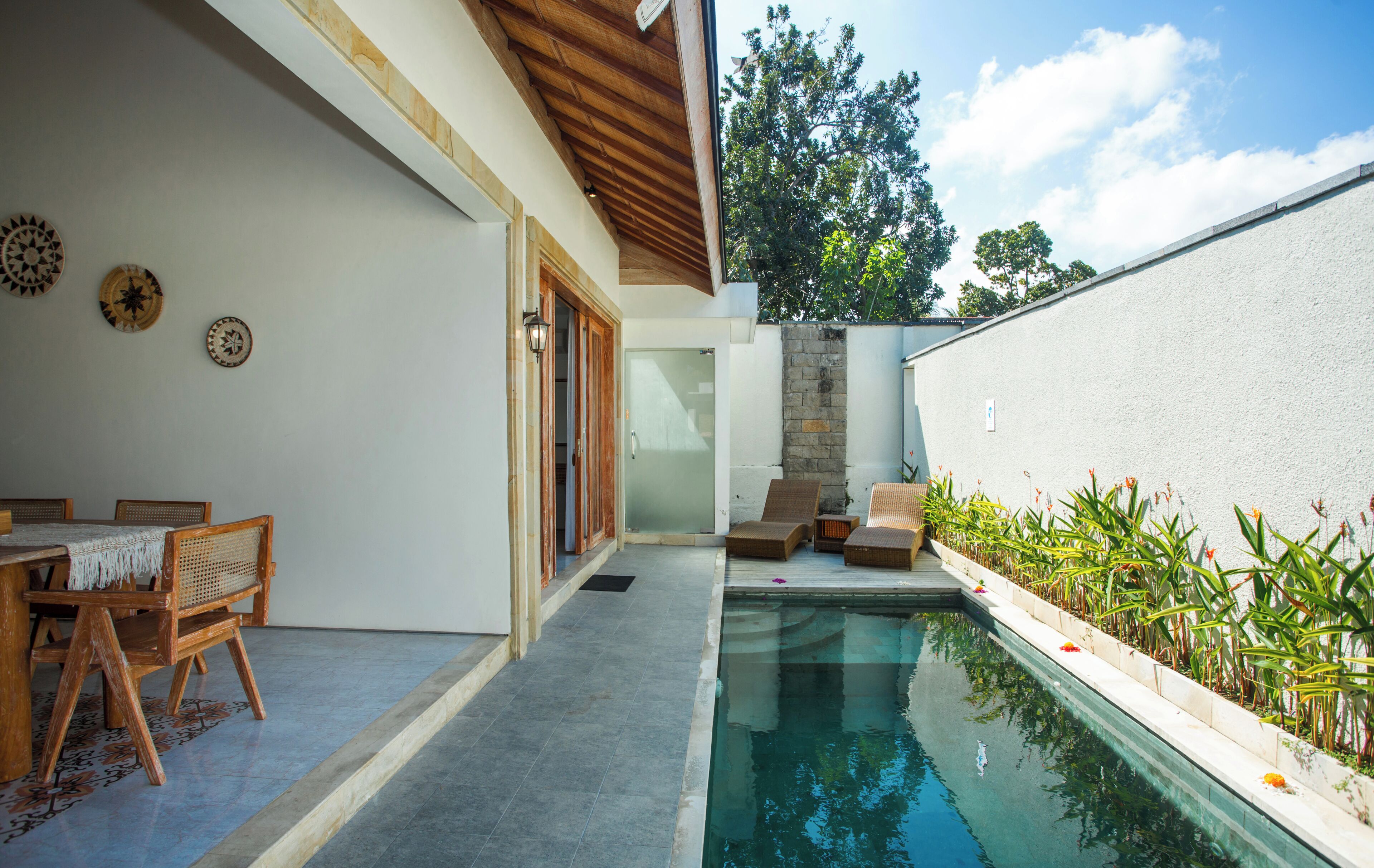 Un área de piscina con mesa y sillas, una pared de piedra y vista a árboles.