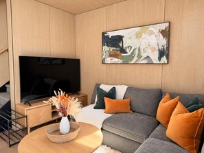 A modern living room with a flat-screen TV, a grey sofa with orange cushions, a wooden coffee table, and a painting on the wall.