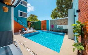 Una piscina esterna moderna con acqua cristallina e blu, circondata da un muro esterno multicolore, una scala in legno e una zona relax con sedie a sdraio.