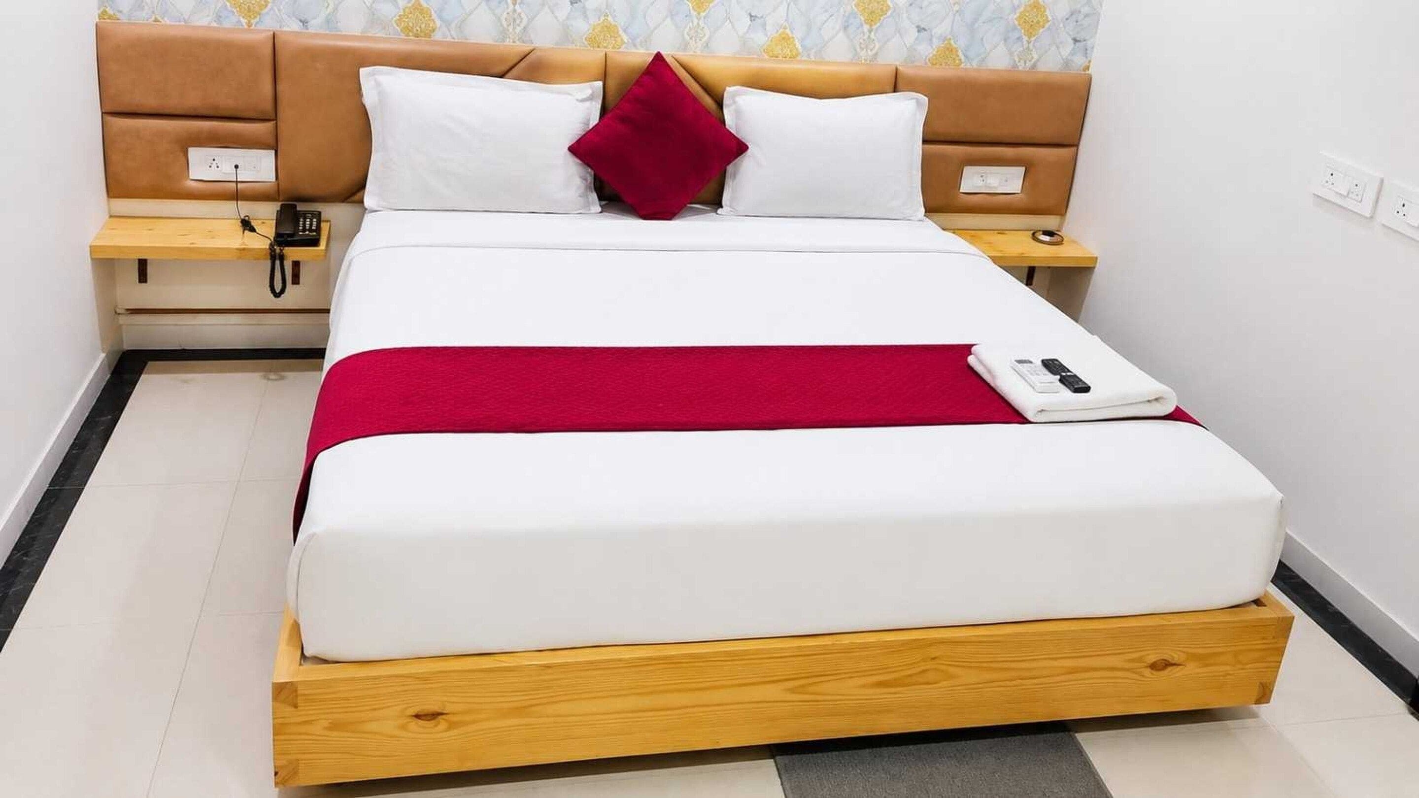 Una cama bien tendida con una manta roja, una mesita de noche con un teléfono y otra mesita con una lámpara.