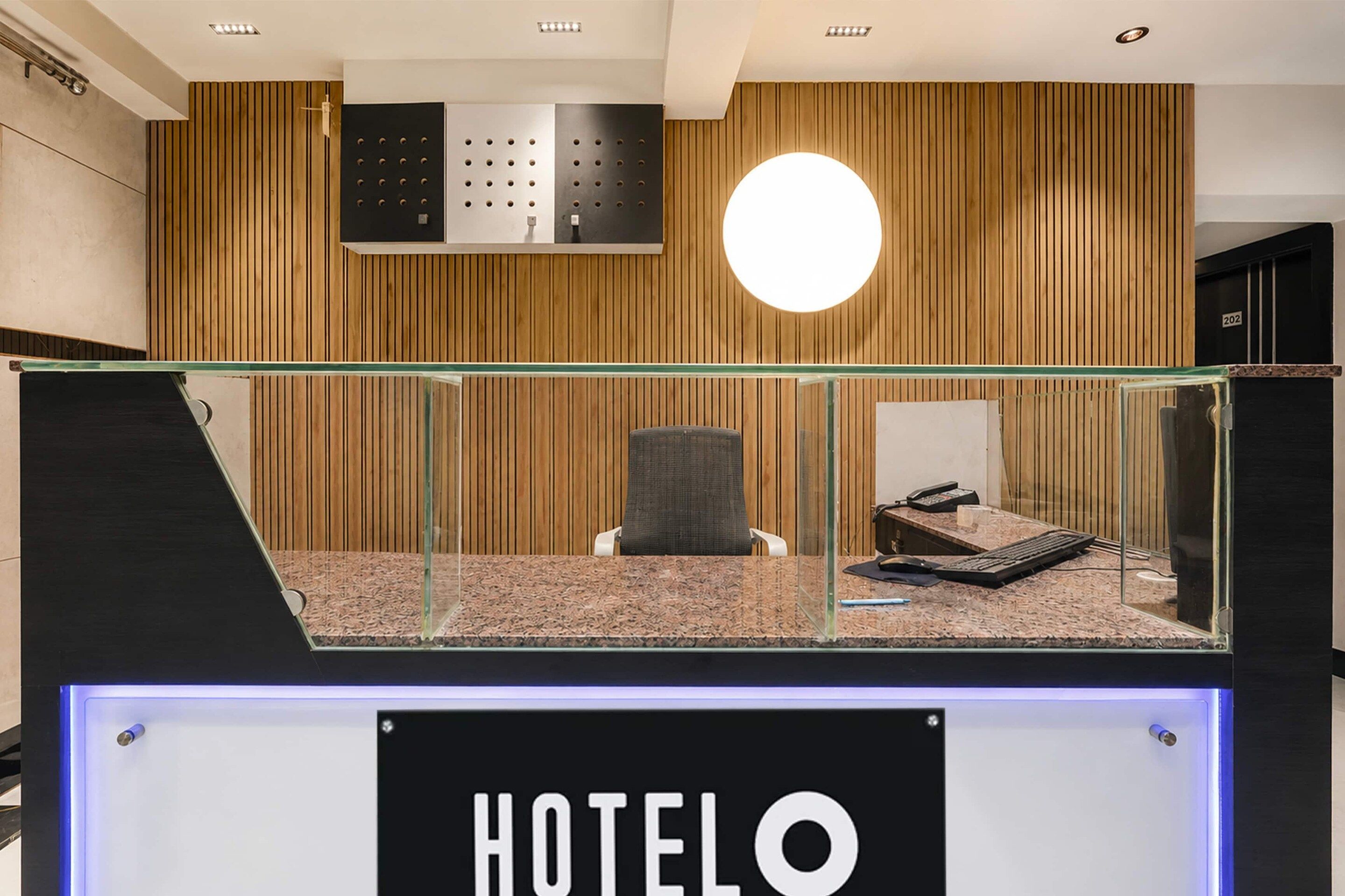A hotel reception desk with a glass partition, a seating area with a chair, and a wooden panel wall with a circular light fixture.
