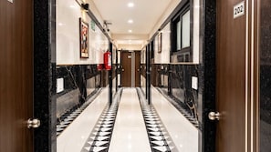 A corridor with a black and white patterned floor, wooden doors, and a fire extinguisher on the wall. - Deluxe Hanspura Bus Terminal (Ahmedabad)