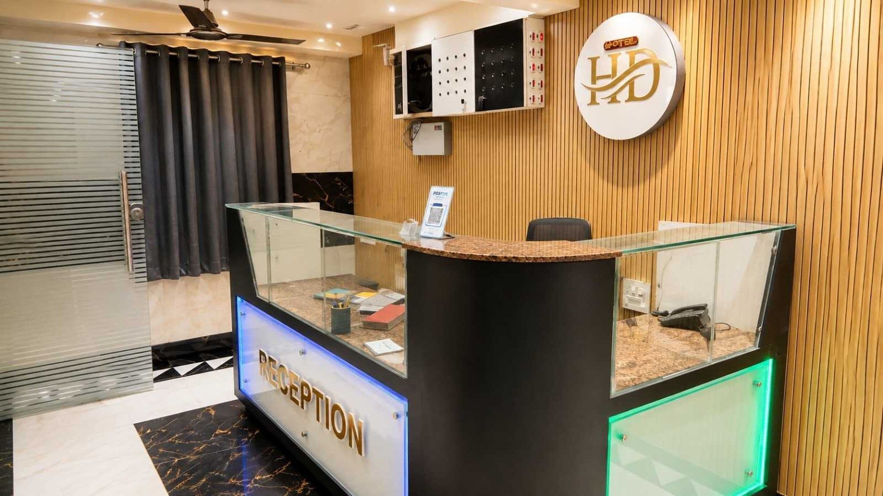A hotel reception area with a glass reception desk, a wooden wall paneling, a ceiling fan, and a sign indicating the hotel's name.