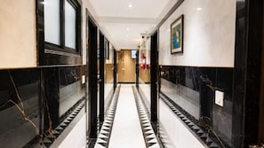 A corridor with black and white patterned flooring, doors on both sides, and a fire extinguisher mounted on the wall. - Deluxe Hanspura Bus Terminal (Ahmedabad)