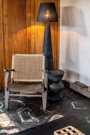 Een houten stoel in rustieke stijl met een geweven zitting, een hoge zwarte lamp en een stenen kom op een stenen vloer.