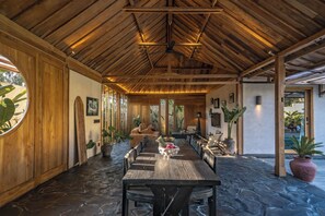A covered outdoor dining area with a stone floor, wooden ceiling beams, and a long table with chairs. - Rahasia Rumah Kerang Uluwatu (Pecatu)