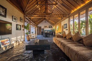 A spacious living area with a high ceiling, a stone floor, a sofa with cushions, a coffee table, and a television. - Rahasia Rumah Kerang Uluwatu (Pecatu)