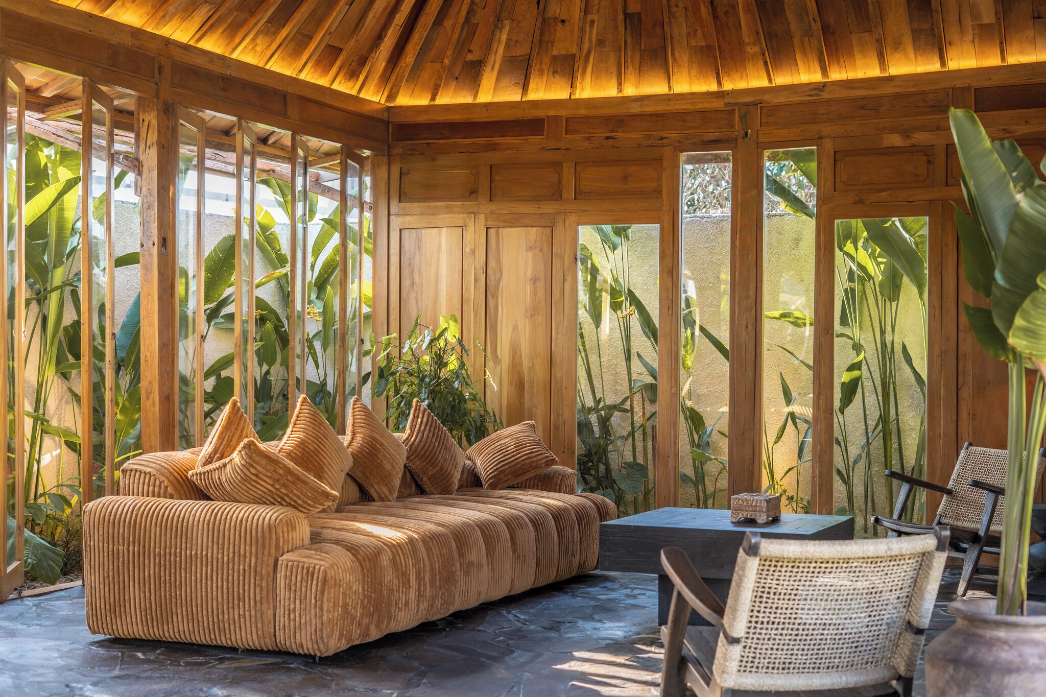 A wooden interior with a sofa and chairs, large windows, and tropical plants.