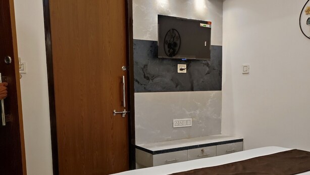 A hotel room with a bed, a wooden door, a flat-screen TV mounted on the wall, and a small table with a power outlet. - Shubh Ashray (UDAIPUR)