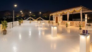 An outdoor terrace with a covered seating area, potted plants, and a clear view of the mountains in the background. - Shubh Ashray (UDAIPUR)