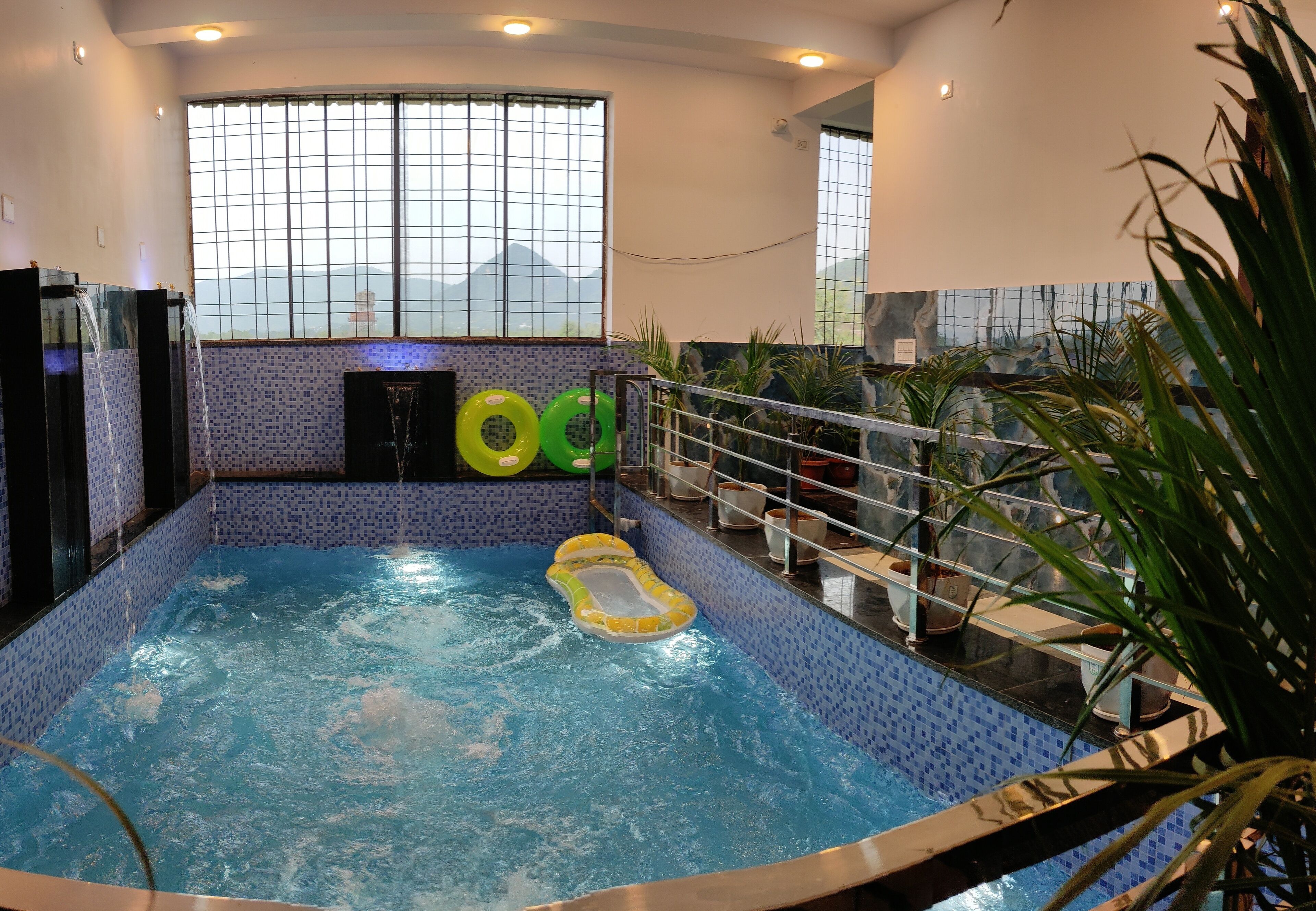 An indoor swimming pool with a view of mountains through the window, surrounded by potted plants.