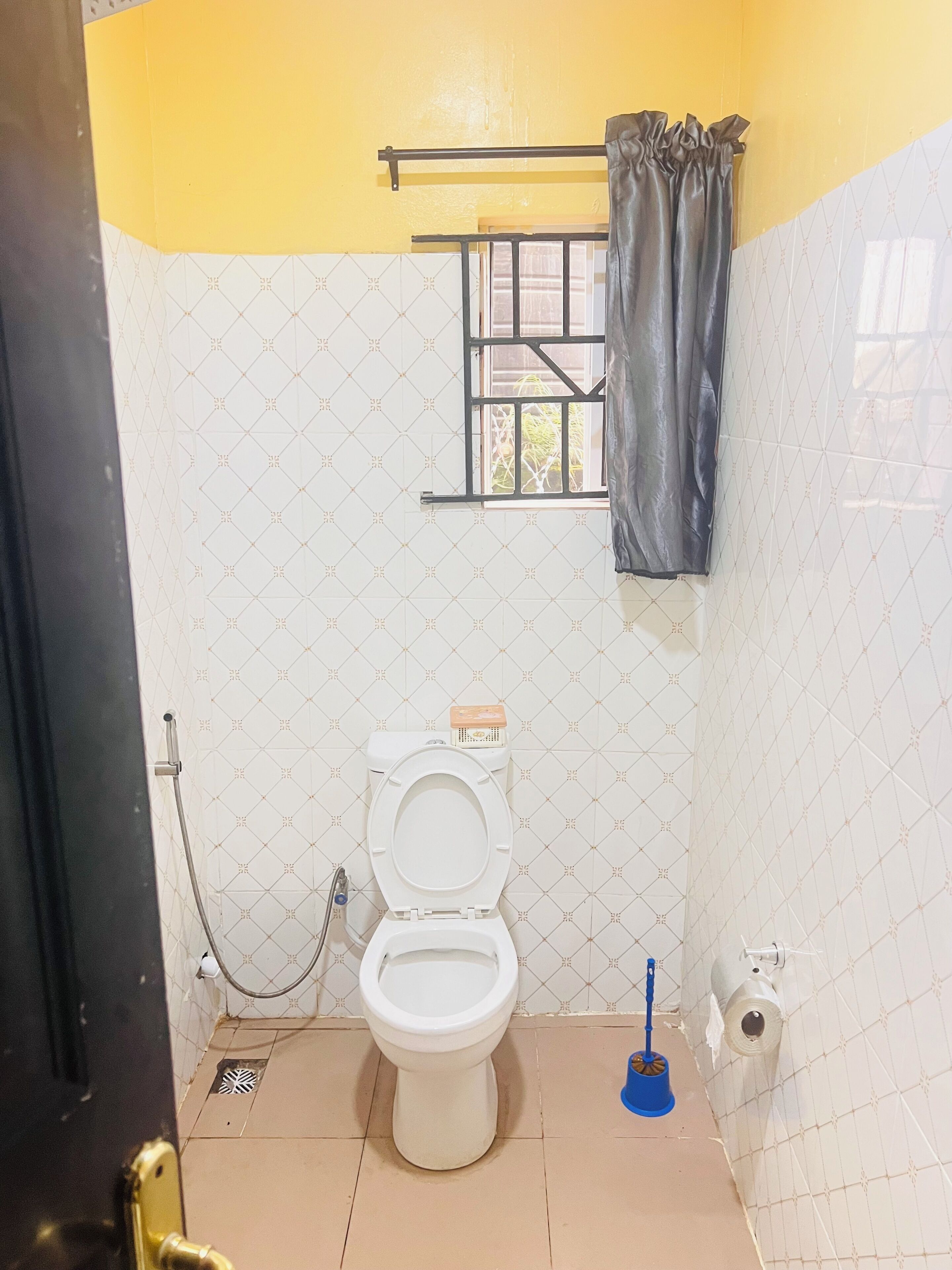 Un bagno piccolo con il water, una finestra con tenda, un tubo flessibile per la doccia e una spazzola blu per le pulizie.