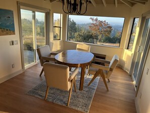 A dining area with a round table and six chairs, large windows with a view of trees and hills, and a chandelier. - Vineyard Hill — Life’s an Adventure. Stay Somewhere Worth the Journey. (Forestville)