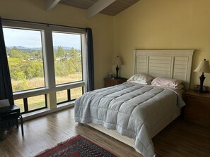 Ein Schlafzimmer mit großem Fenster, einem Bett, einem Nachttisch und einer Lampe.