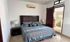 Quarto com cama, mesas de cabeceira, ar condicionado fixado na parede e porta deslizante para um banheiro.