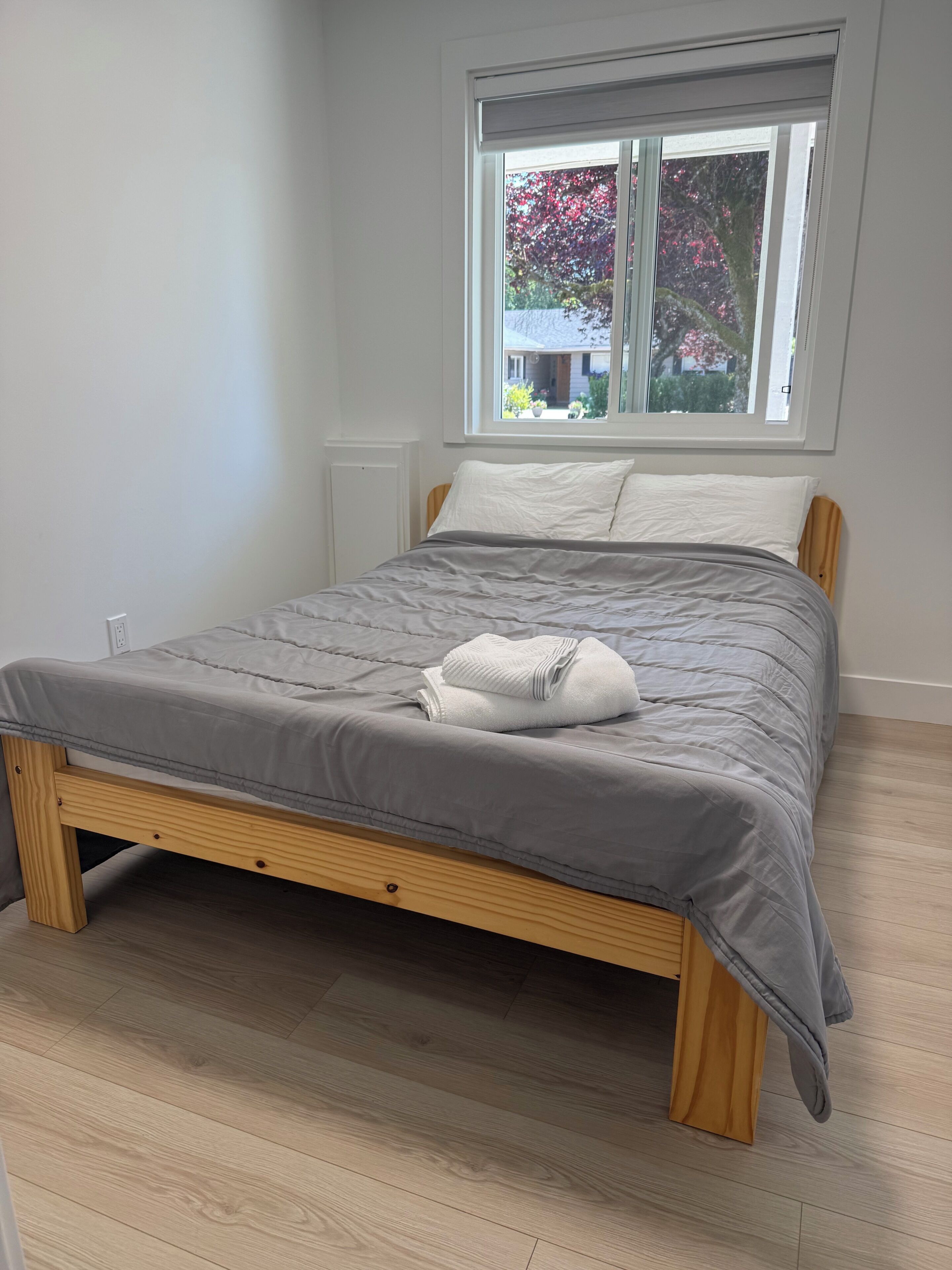 Una cama con edredón gris, estructura de madera y ventana con vistas a zonas verdes.
