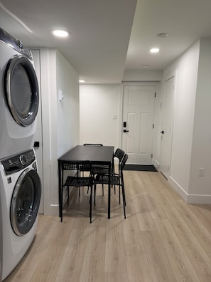 Uma lavanderia com máquina de lavar, mesa e cadeiras, e uma porta ao fundo.