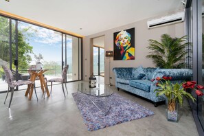 A modern living room with a blue sofa, a glass coffee table, and a large window offering a view of greenery. - Art Deco 3 BR Pool Villa Kamala NP1 (Kathu)
