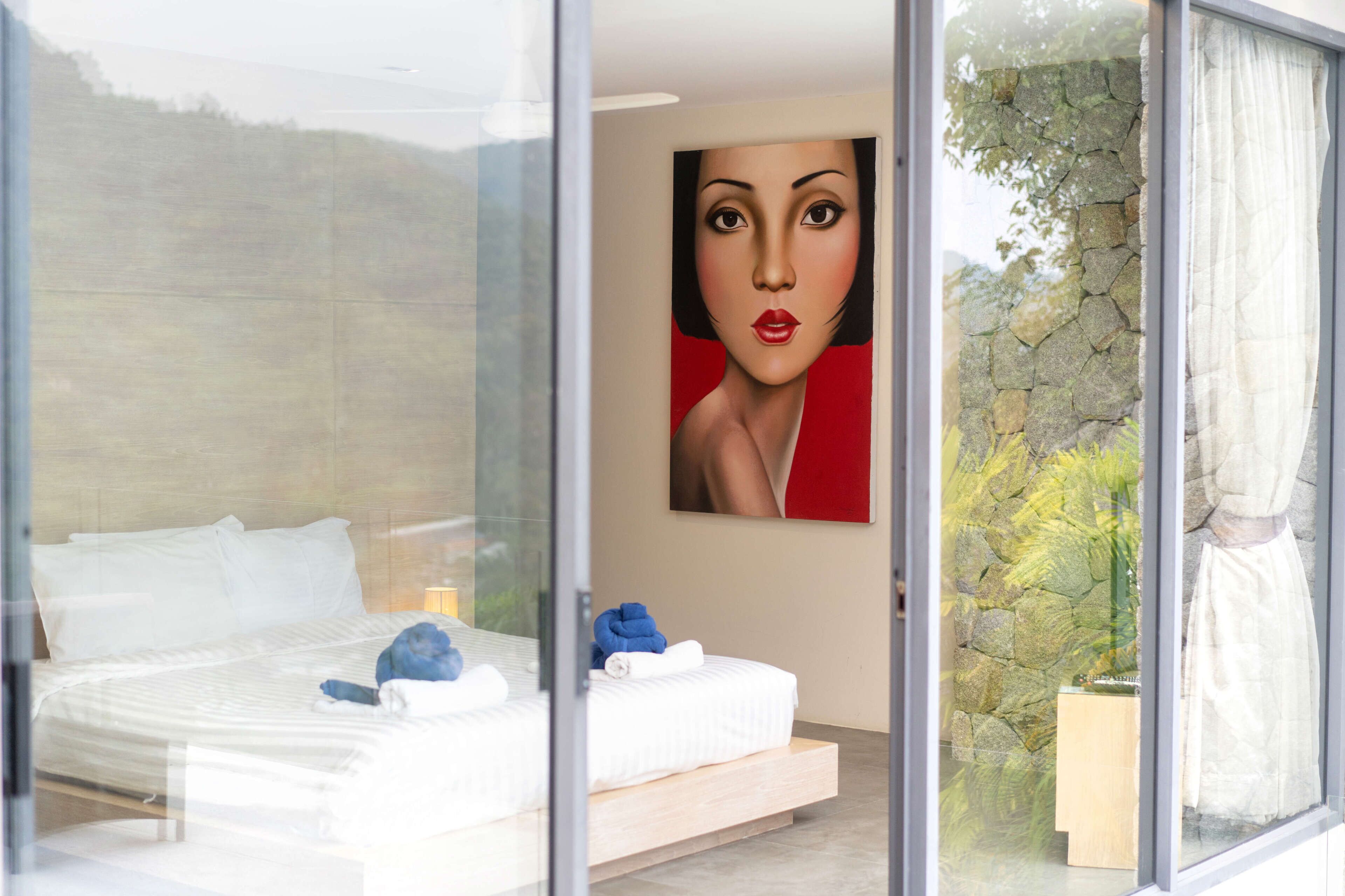 Un dormitorio moderno con pared de vidrio, cama con ropa blanca, toalla azul y pared de piedra con vegetación afuera.