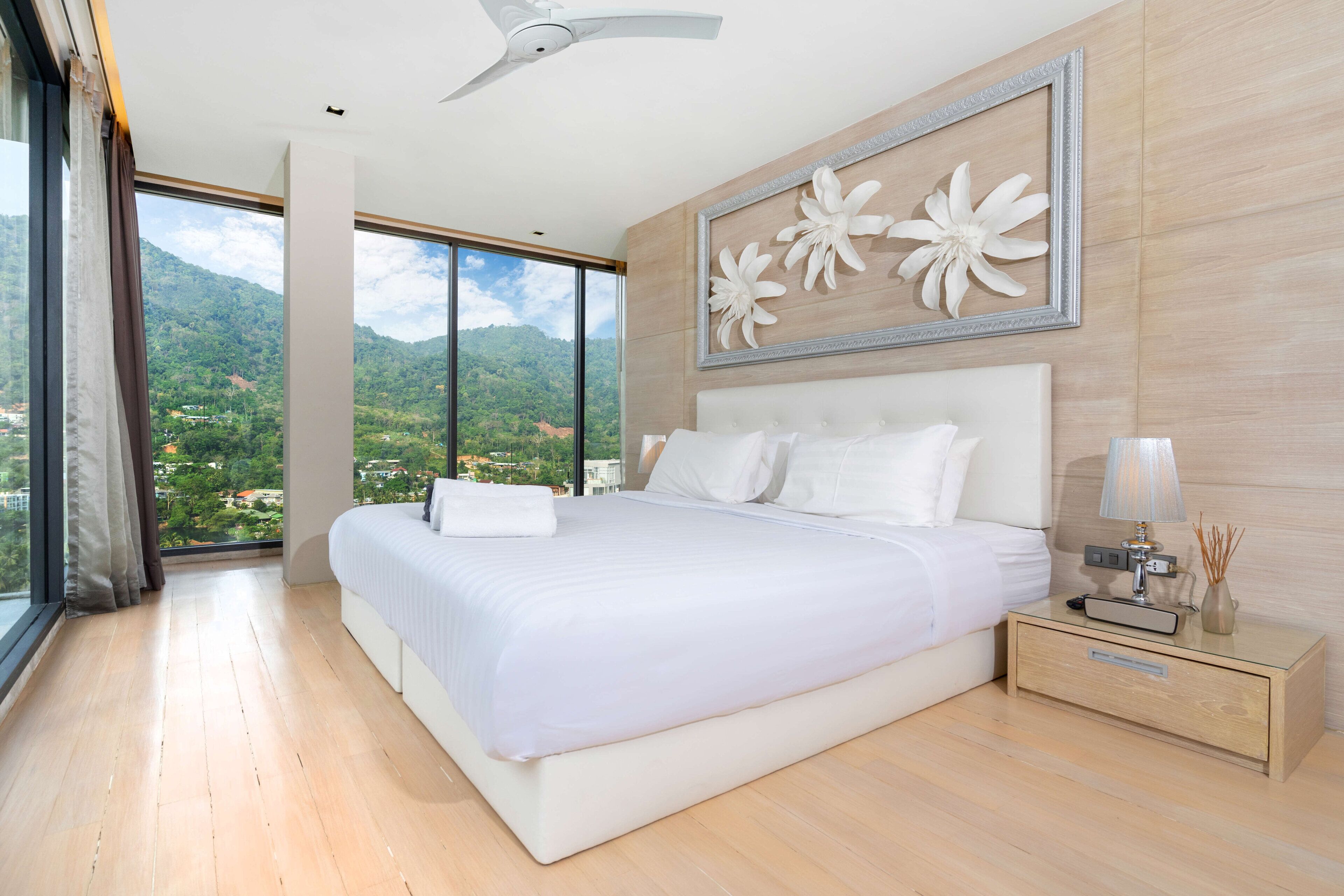 Un dormitorio moderno con una cama grande, una mesita de noche, una lámpara y vistas a un paisaje montañoso a través de amplios ventanales.