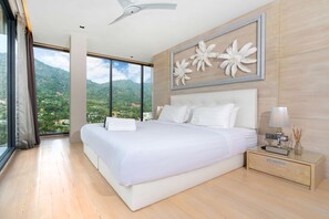 A modern bedroom with a large bed, a nightstand, a lamp, and a view of a mountainous landscape through large windows. - Art Deco 3 BR Pool Villa Kamala NP1 (Kathu)