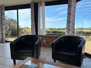 A room with two black leather chairs, a glass-top table, and a view of a green landscape through large windows. - Relaxing Holiday Home in Normandy (Courtils)