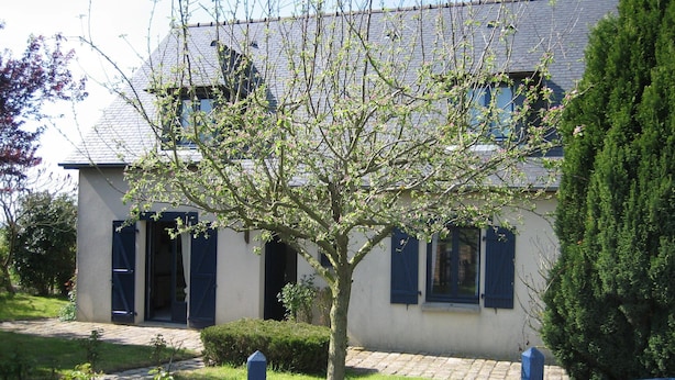 Ein zweistöckiges Haus mit Baum im Vorgarten, einem gepflasterten Einfahrtsweg und einem blauen Zaun.