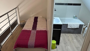 Ein kleines Schlafzimmer mit einem Bett, einem Nachttisch, einer grünen Mülleimer und einem Badezimmer mit Waschbecken und Badewanne.