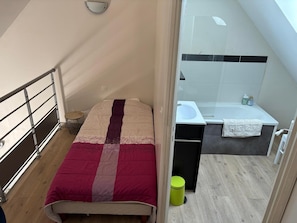A small bedroom with a bed, a bedside table, a green trash bin, and a bathroom with a sink and bathtub. - Relaxing Holiday Home in Normandy (Courtils)