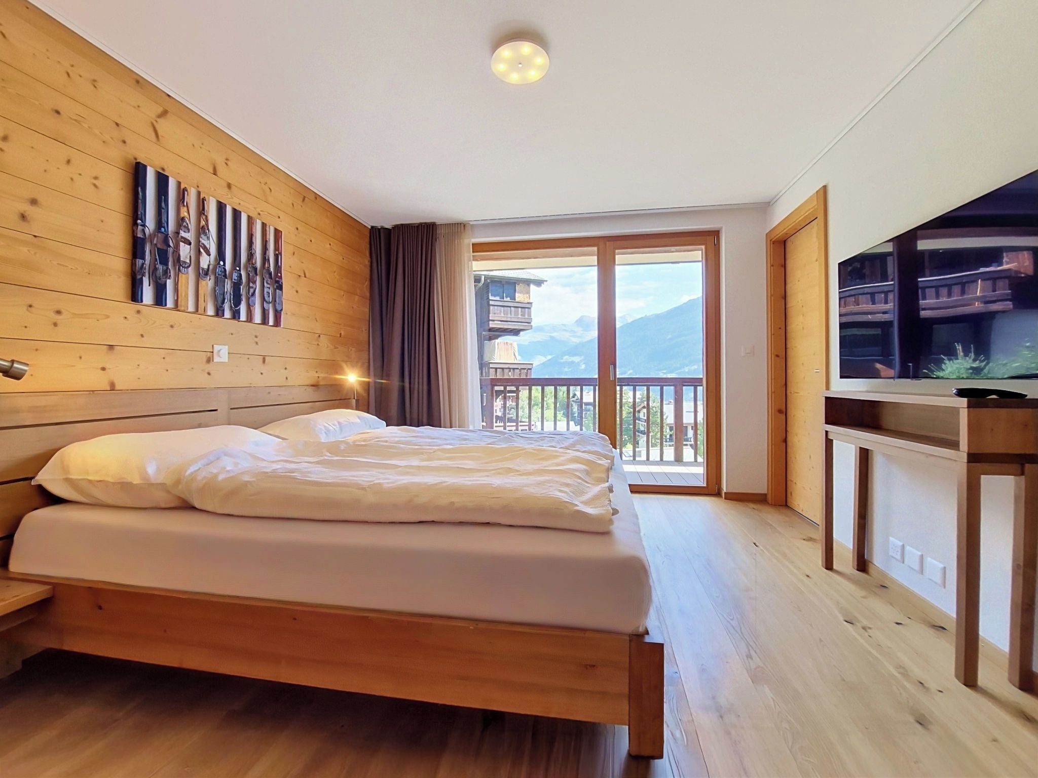 A bedroom with a wooden headboard, a bed with white bedding, a large window with a view of mountains, and a wooden TV stand.
