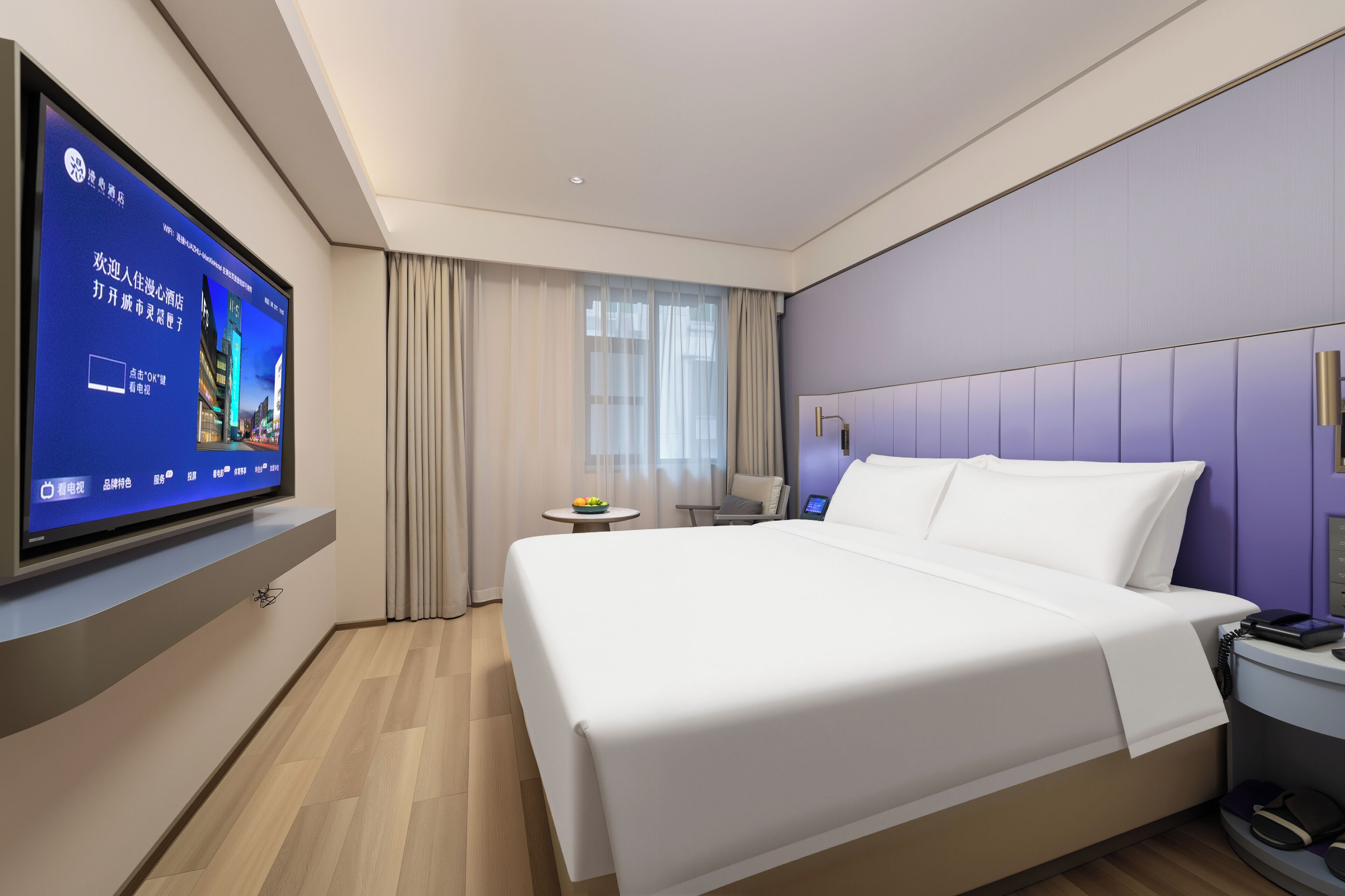 Ein modernes Hotelzimmer mit einem großen Bett, einem flachbildfernseher an der Wand, einem kleinen Tisch mit Obstschale und einem Nachttisch mit Telefon.