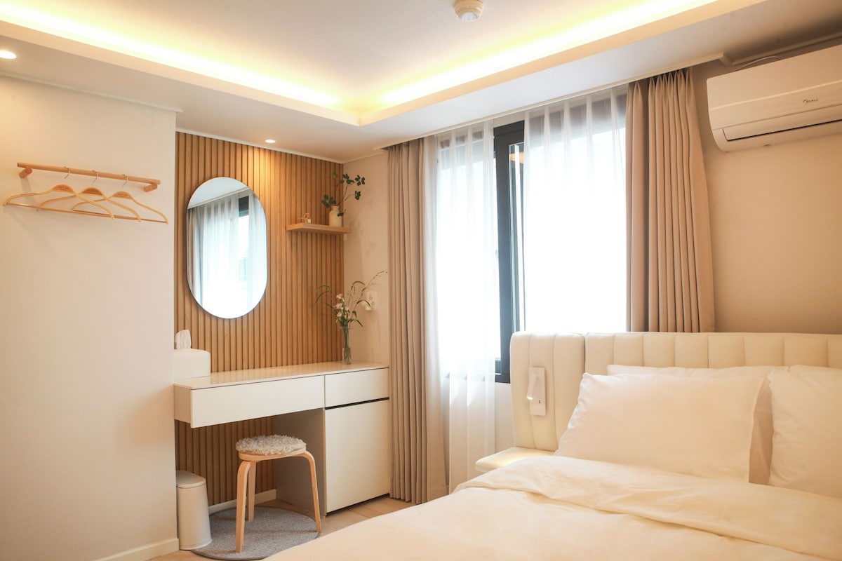 A hotel room with a bed, a desk with a chair, a mirror, a small stool, a plant, and a window with curtains.