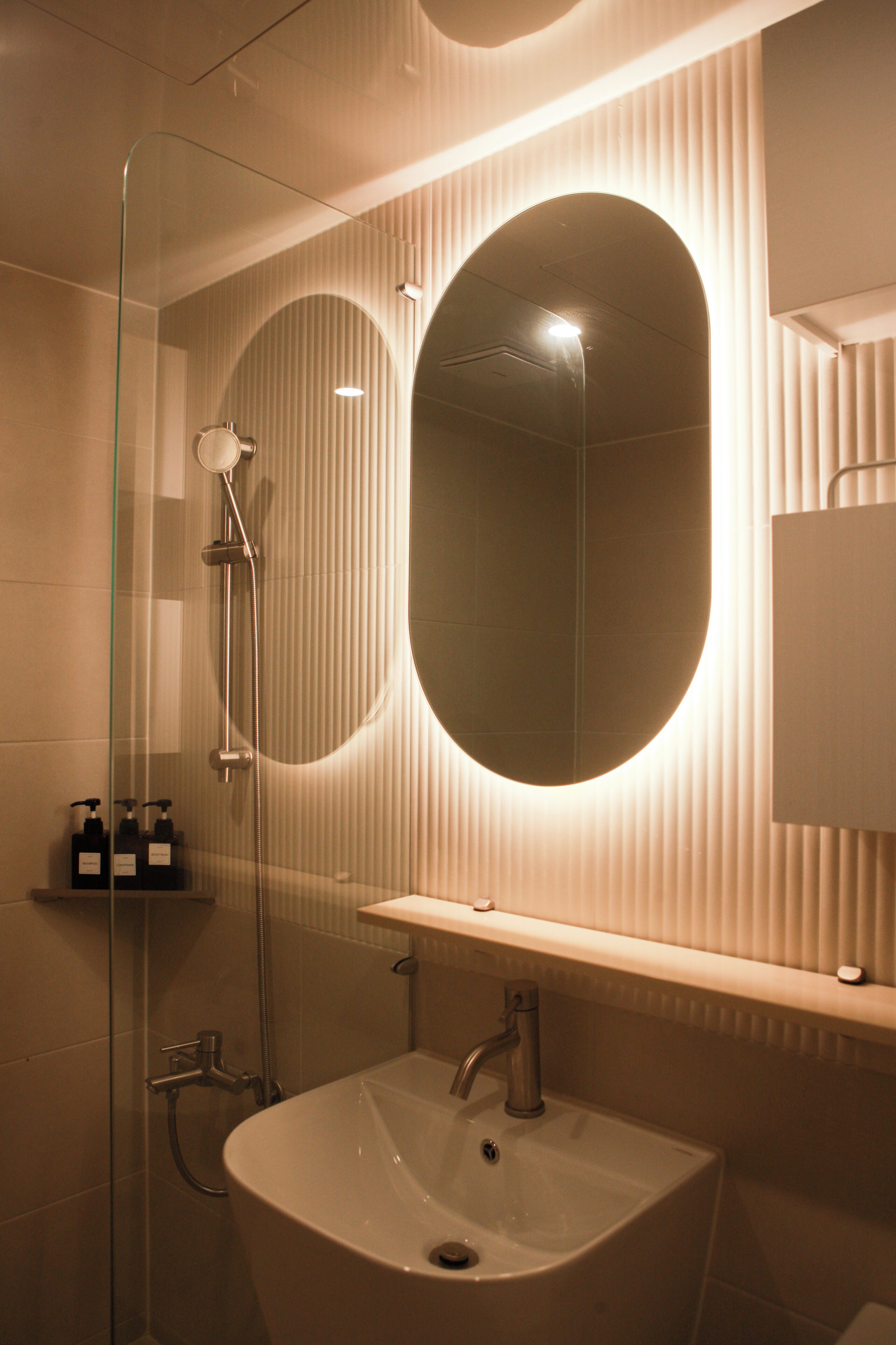 Un baño moderno con un espejo ovalado grande, ducha con mampara de vidrio y lavamanos blanco con un solo grifo.