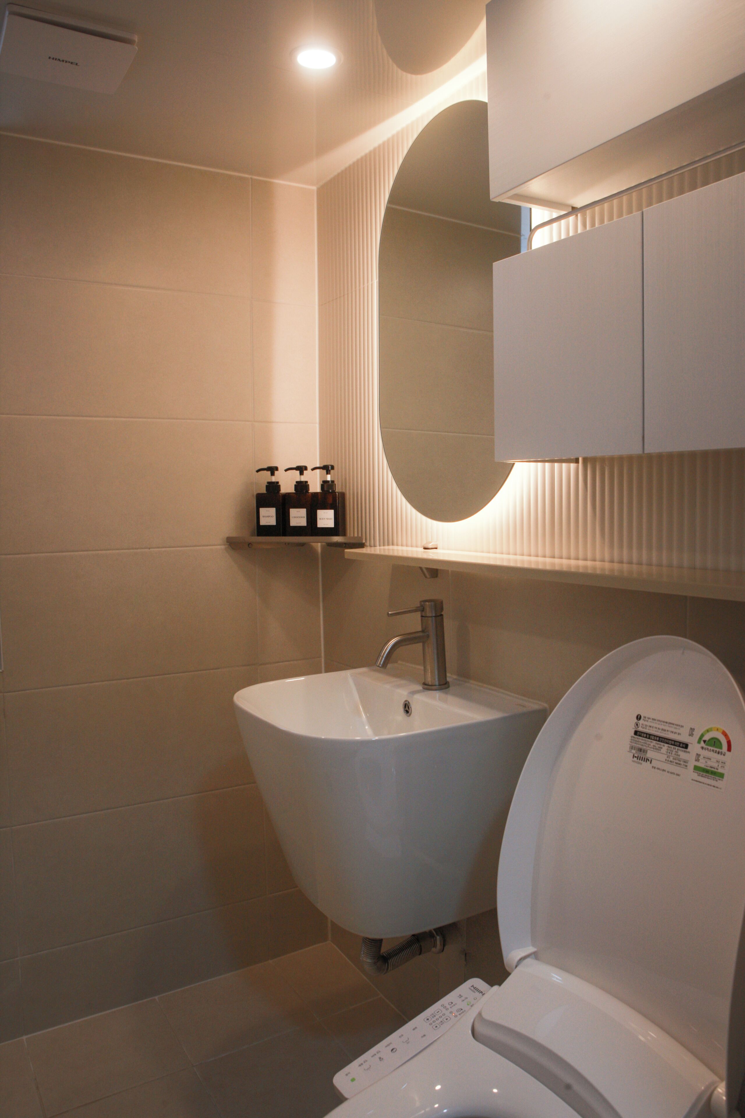 A bathroom with a white sink, a toilet, and a mirror. There are three bottles on the wall.