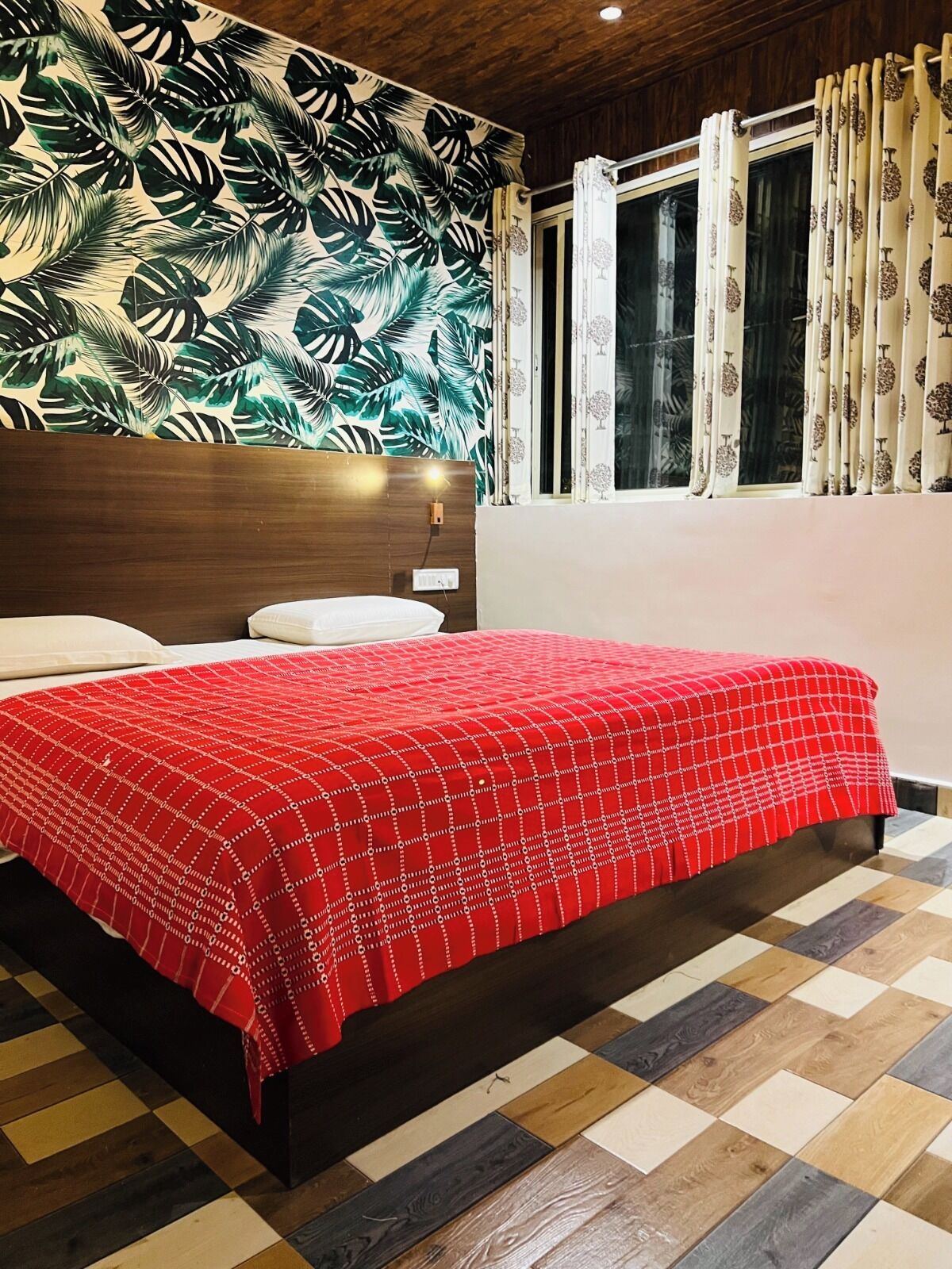 Un dormitorio con pared estampada, cama con colcha roja, ventana con cortinas y cabecera de madera.