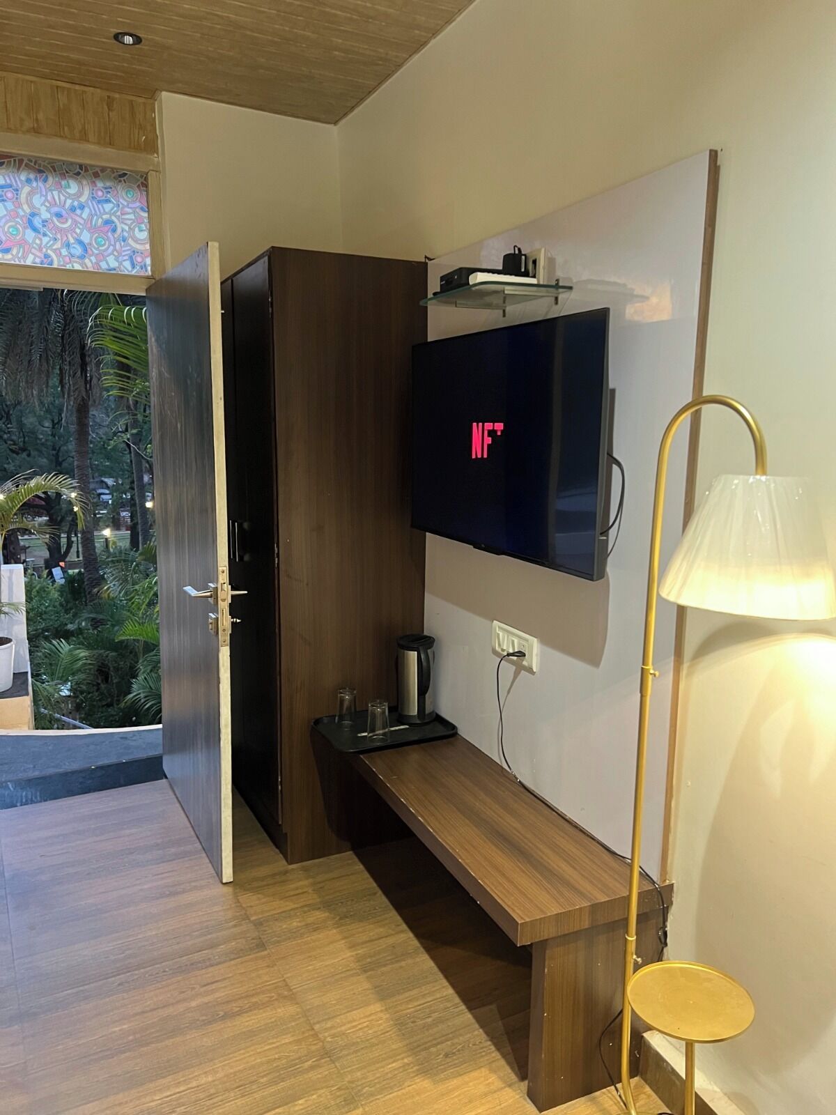 Una habitación moderna con piso de madera, televisor montado en la pared, un banco, una lámpara y una puerta que da a un espacio exterior con vegetación.