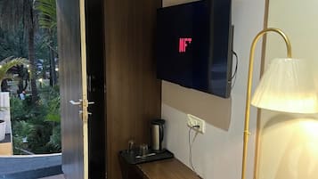 Una habitación moderna con piso de madera, televisor montado en la pared, un banco, una lámpara y una puerta que da a un espacio exterior con vegetación.