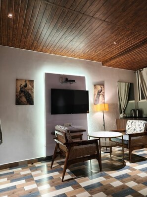 A room with a flat-screen TV mounted on the wall, a round table with chairs, a lamp, and artwork depicting peacocks. - Hotel Rudra Villa (Abu Road)