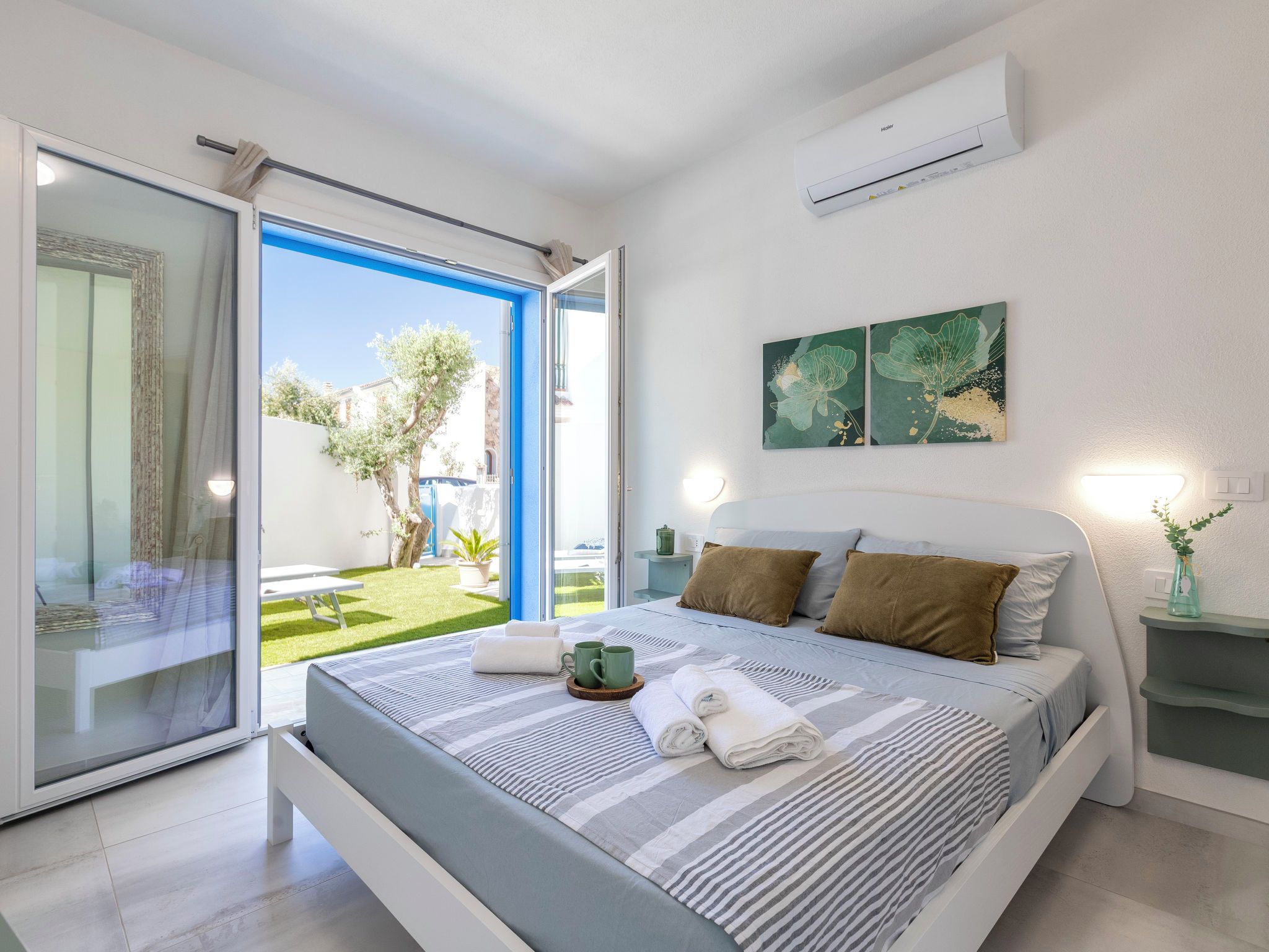 A modern bedroom with a bed, pillows, a nightstand, a wall-mounted air conditioner, and a view of a courtyard.