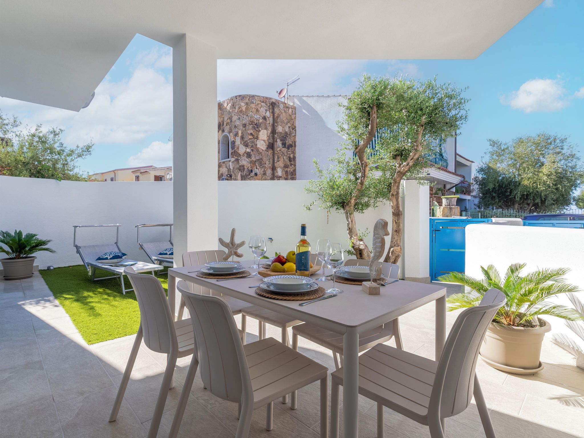 Un espacio de comedor al aire libre con una mesa dispuesta para dos personas, rodeado de sillas, un pequeño jardín y vistas a un muro de piedra y árboles.